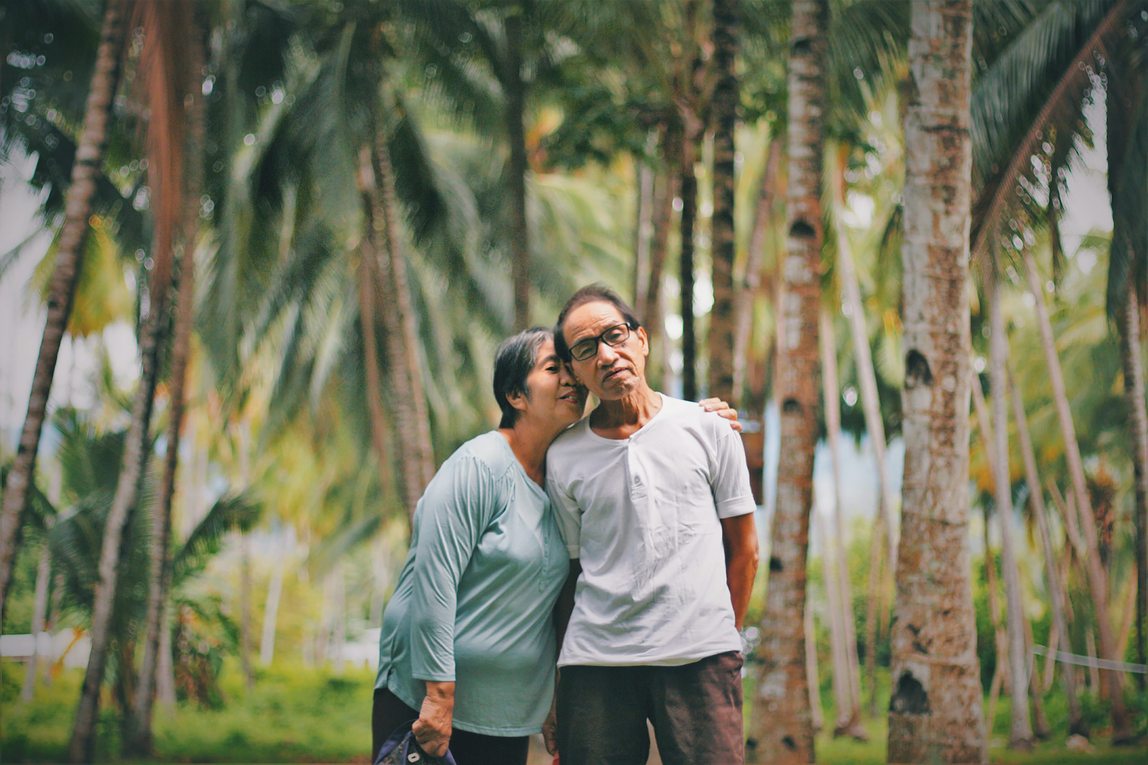 elderly woman putting arm around husband standing among tropical trees