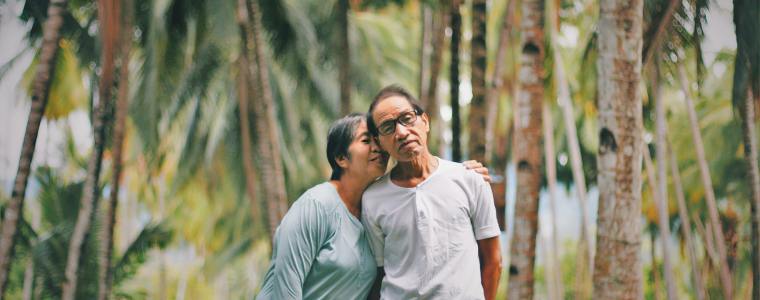elderly woman putting arm around husband standing among tropical trees